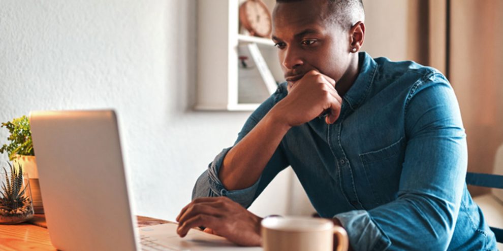 Black man reading laptop with cup of coffee.